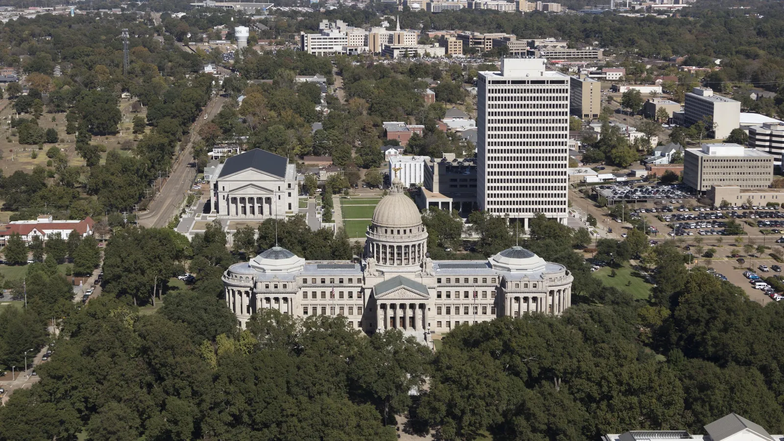 Aerial view of the Mississippi State Capitol in Jackson, a pale stone Beaux-Arts building with a central dome, ringed by trees, with office towers behind it.
